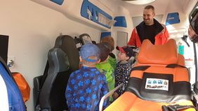 Kinder schauen neugierig auf die Geräte in einem Rettungswagen, ein Notfallsanitäter sieht ihnen dabei zu.