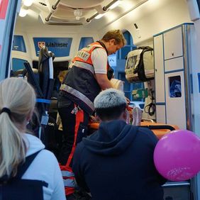 Ein junger Mann in Sanitäterkleidung zeigt interessierten Menschen einen Rettungswagen von innen.